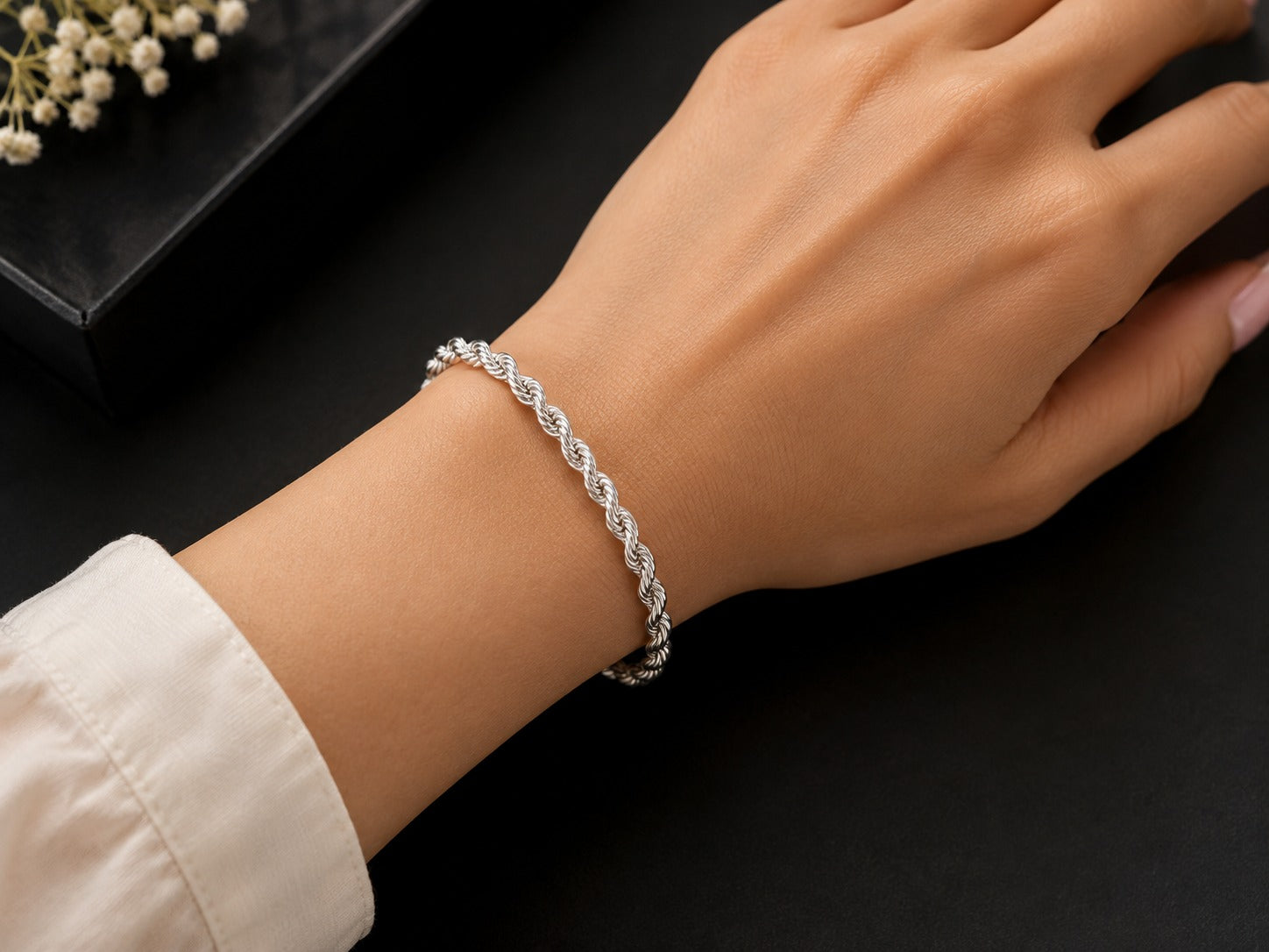 A silver twisted rope bracelet worn on a woman's wrist, shown against a dark background with a black box and small white flowers as props.