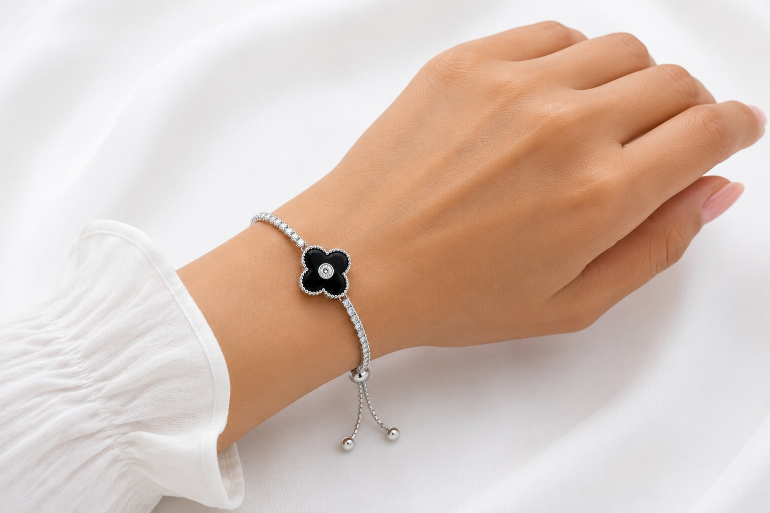 A delicate silver-toned bracelet with a black flower-shaped centerpiece and a small clear stone, shown worn on a person's wrist against a soft white fabric background.