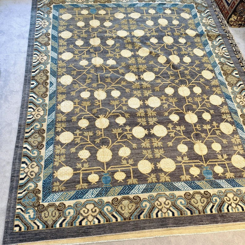 Afghan hand-knotted Khotan wool rug with charcoal grey field, cream pomegranate tree motifs, and blue geometric borders.