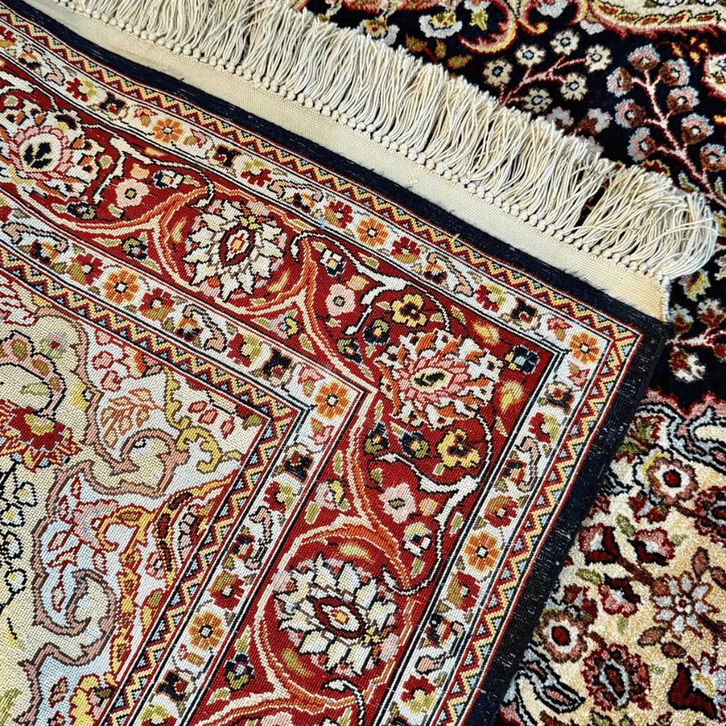 Detailed view of the Kashmiri silk rug's decorative red, cream, and gold floral border against a dark navy background.